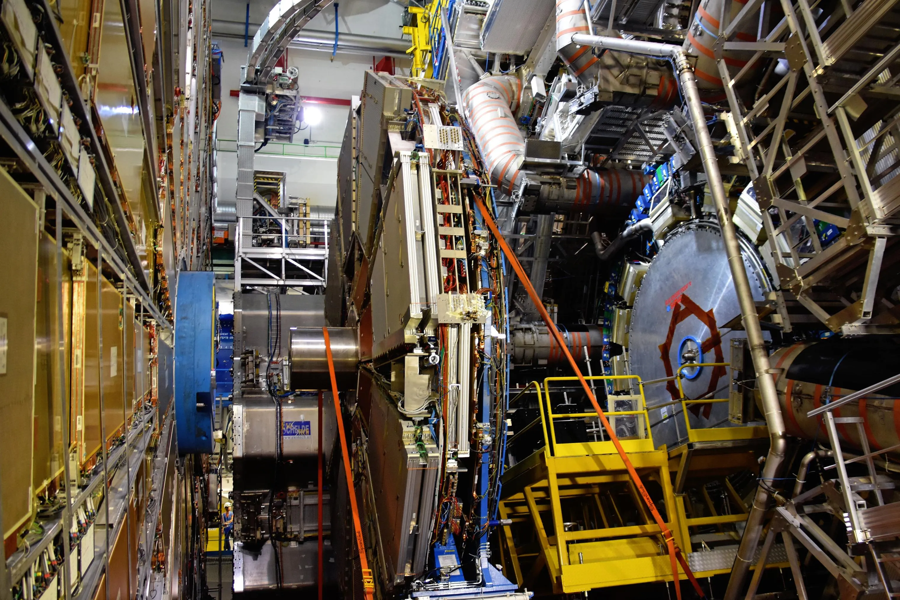 Vista interior del detector ATLAS del LHC, con estructuras metálicas, cables de colores, plataformas amarillas con barandillas y una cinta naranja en diagonal, entre andamios y maquinaria industrial.