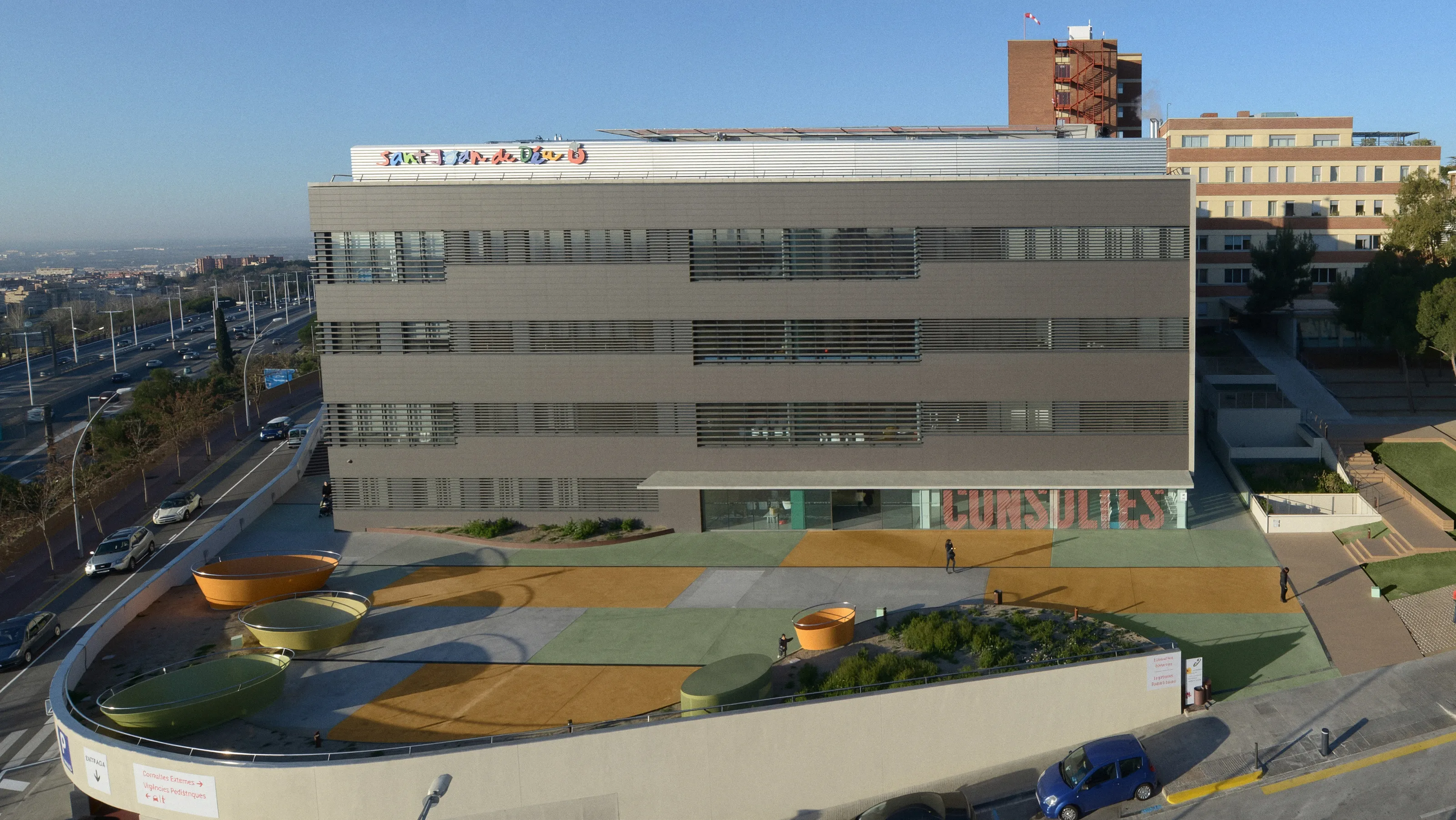 Vista elevada del edificio de consultas externas del Hospital Sant Joan de Déu Barcelona, con el rótulo de colores del hospital en lo alto y una plaza de pavimento verde, ocre y gris con jardineras ovaladas delante.