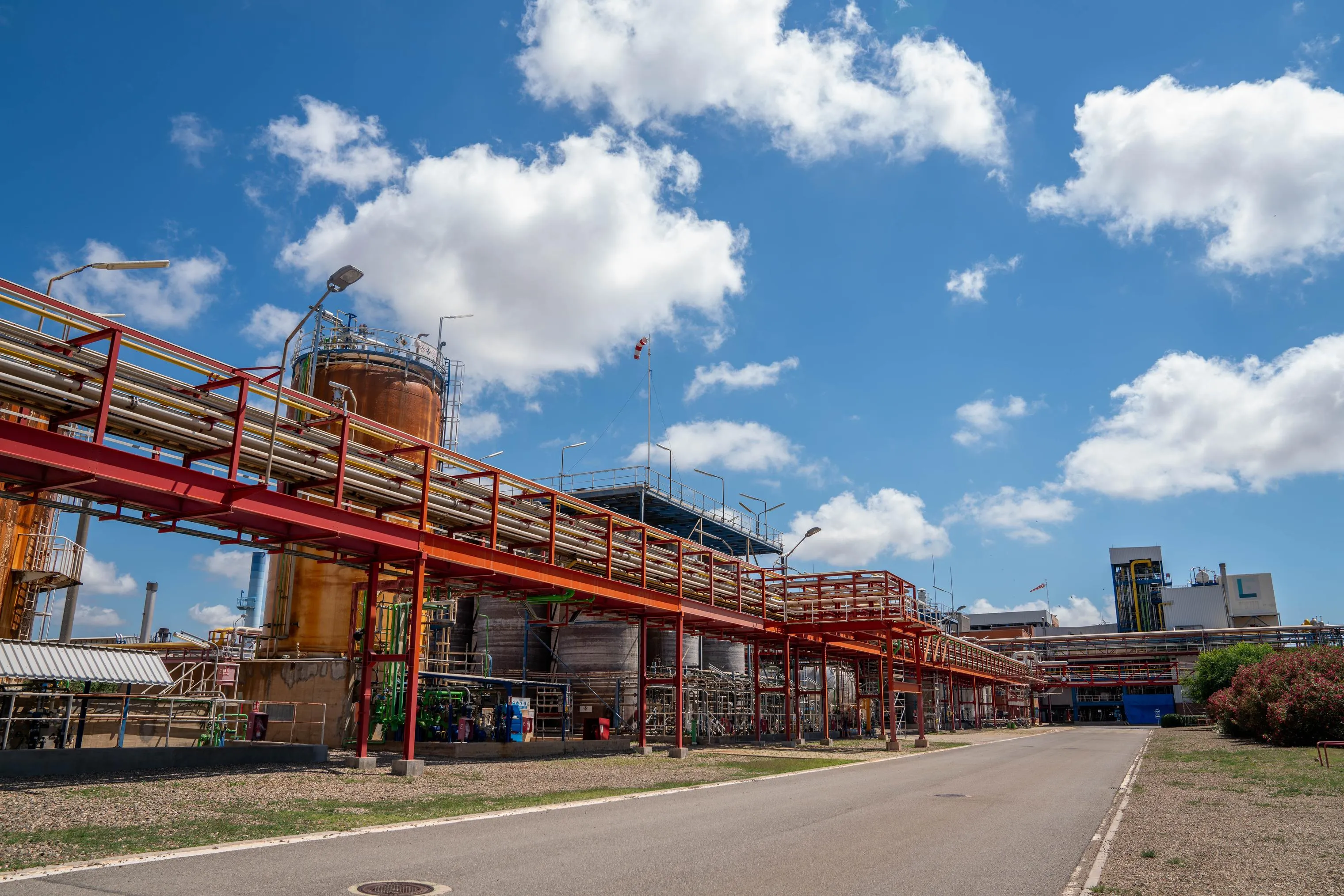 Vista de una planta petroquímica con pasarelas y tuberías metálicas elevadas de color rojo, tanques industriales y un cielo azul con nubes.