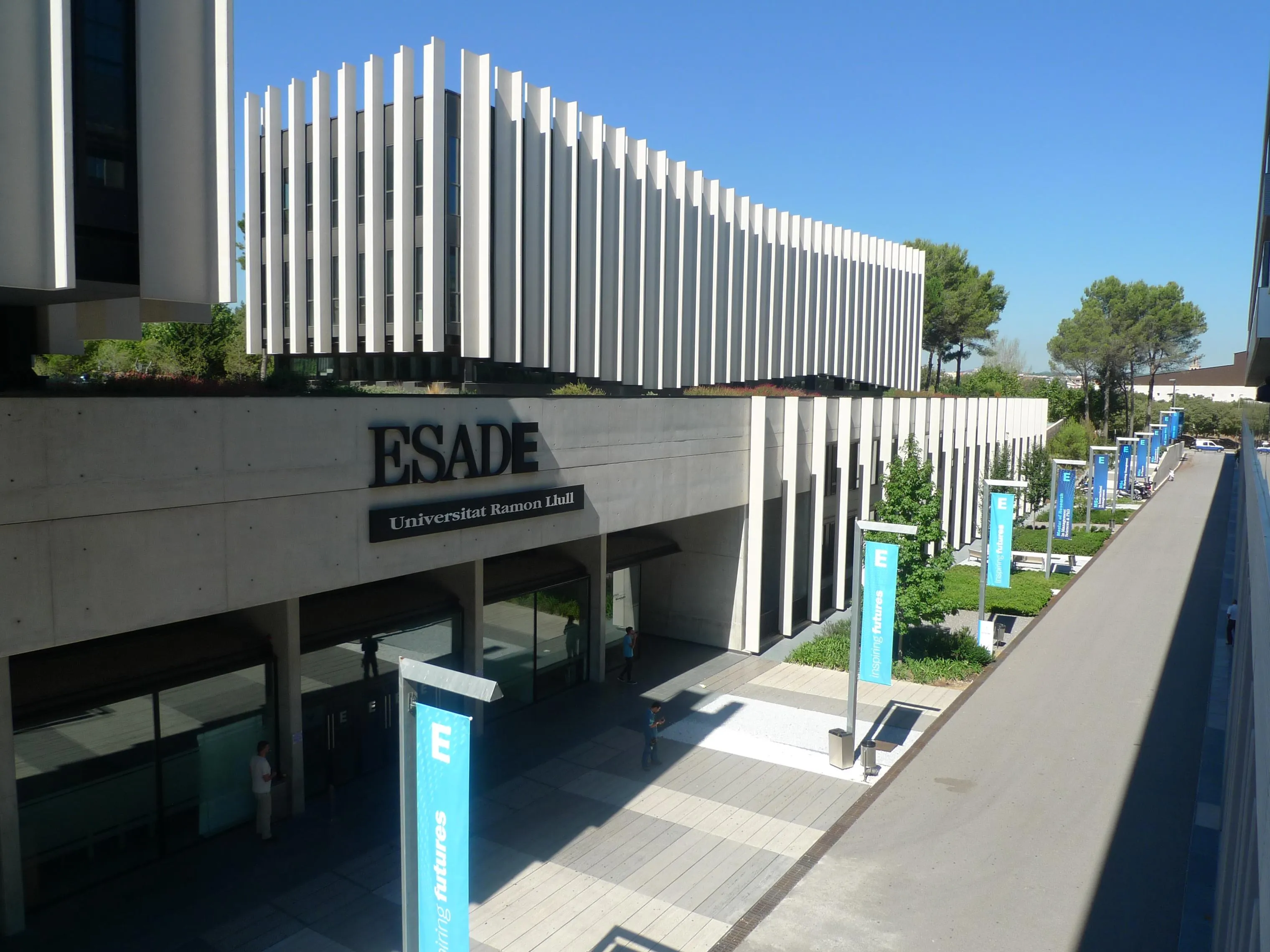 Fachada del campus de ESADE con el rótulo «ESADE, Universitat Ramon Llull», pancartas azules con el lema «Inspiring futures» y varias personas caminando por la entrada.