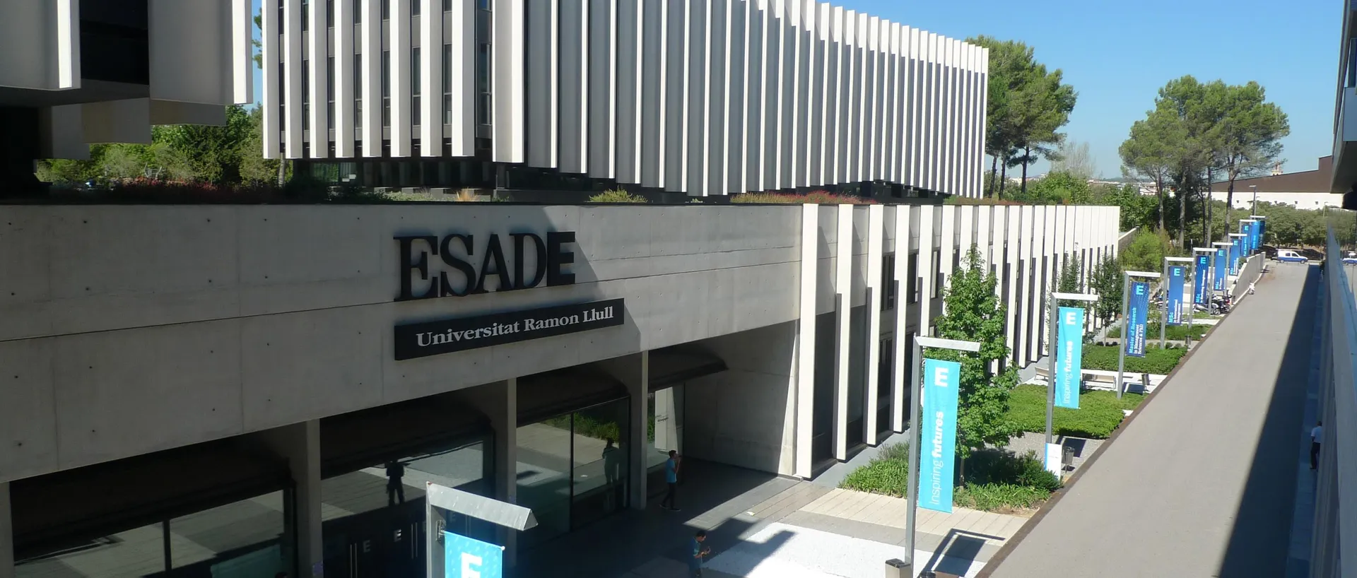 Fachada del campus de ESADE con el rótulo «ESADE, Universitat Ramon Llull», pancartas azules con el lema «Inspiring futures» y varias personas caminando por la entrada.