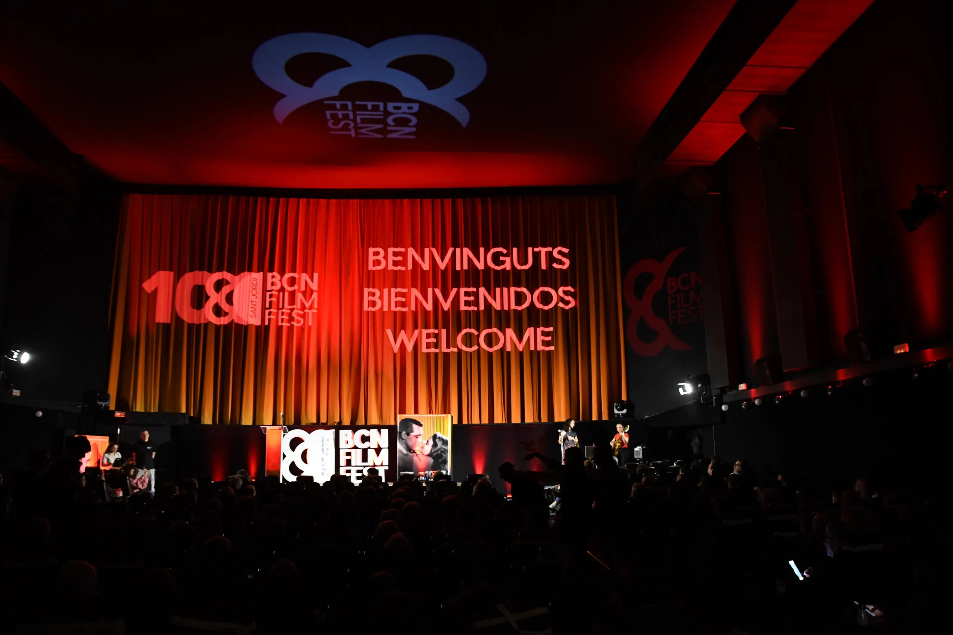 Interior de una sala de cine llena de público, con la pantalla iluminada en rojo mostrando el logo «10&BCN Film Fest» y el texto «Benvinguts, Bienvenidos, Welcome», y dos músicos en el escenario.