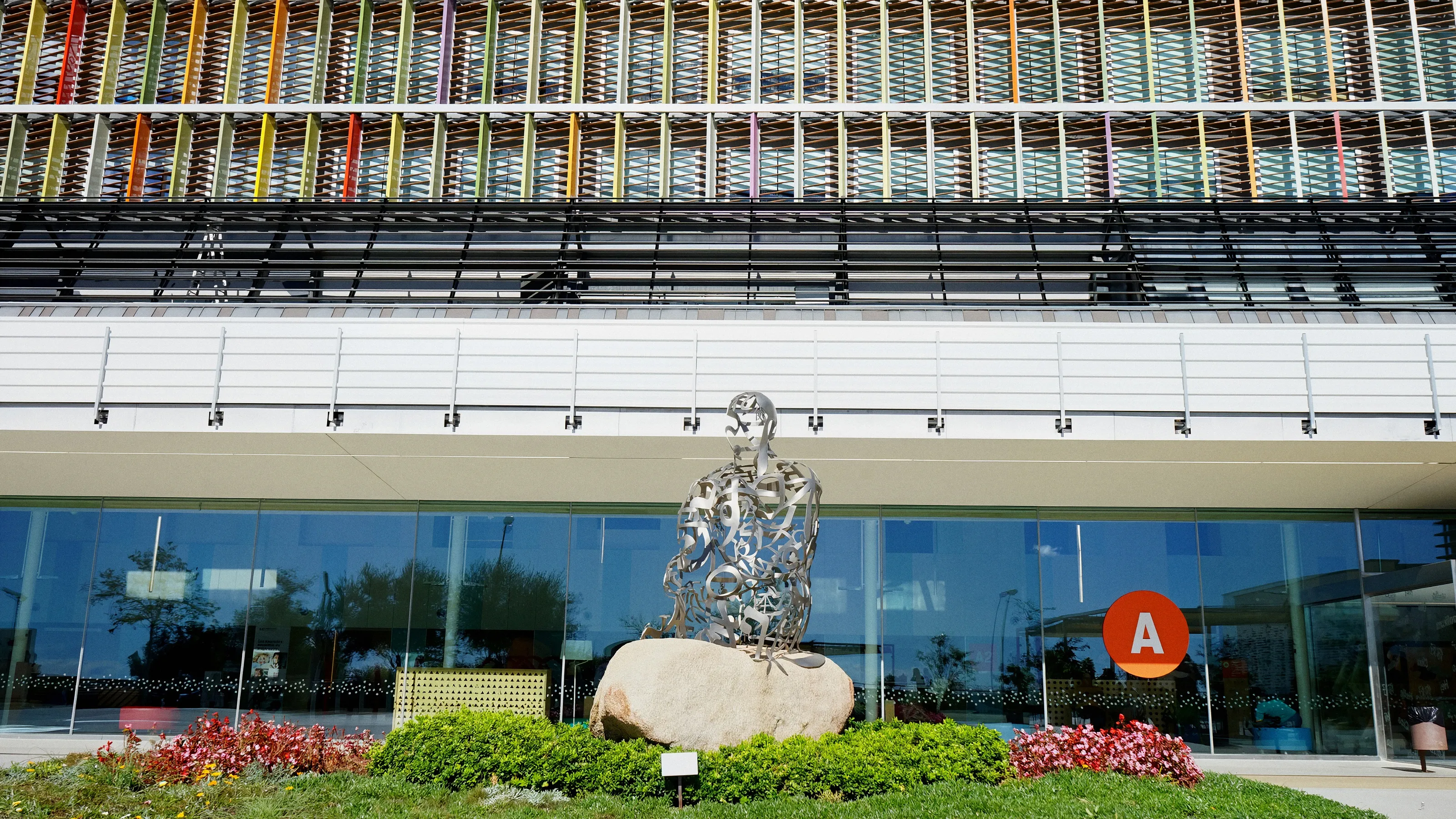 Escultura de Jaume Plensa, una figura sentada hecha de letras de acero sobre una roca, ante la fachada acristalada del Hospital Sant Joan de Déu Barcelona, con lamas de colores en las plantas superiores.