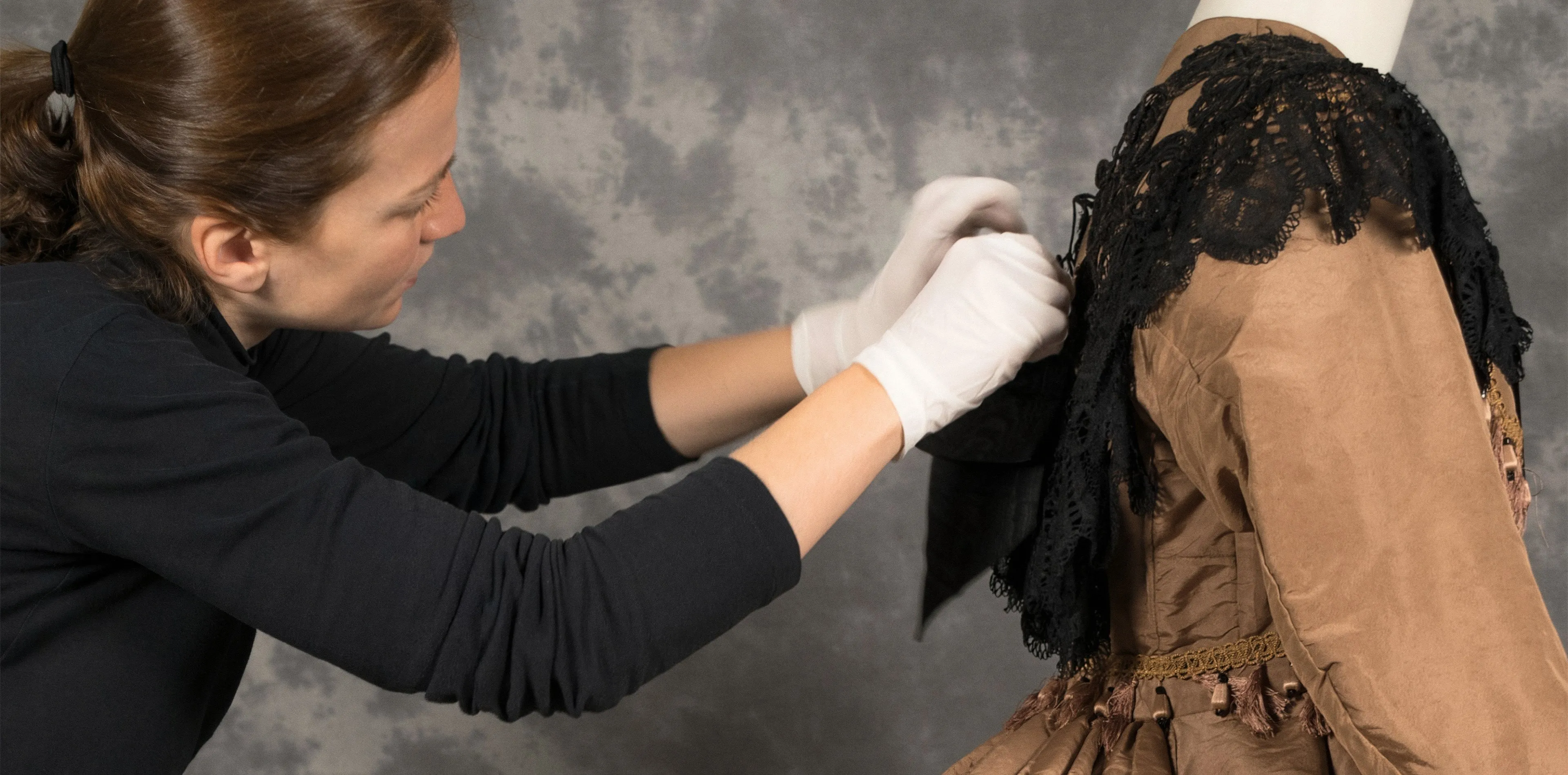 Una restauradora con guantes blancos ajusta el cuello de encaje negro de un vestido histórico de seda marrón colocado en un maniquí.