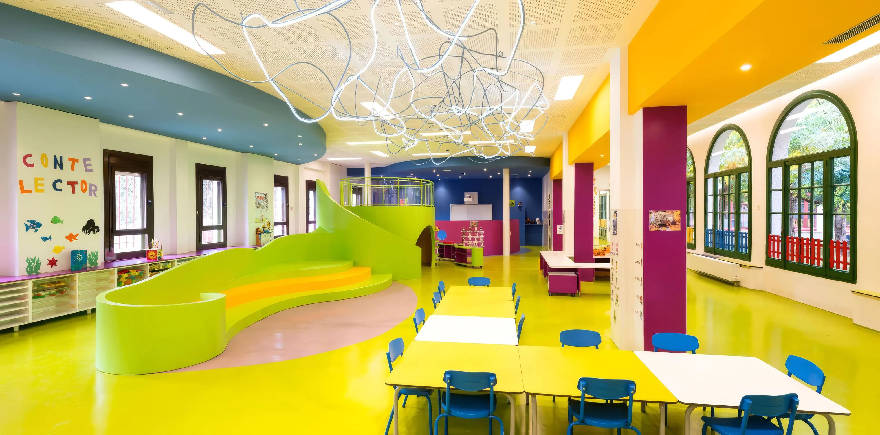 Espacio infantil de un colegio, sin personas: mesas amarillas con sillas azules, una grada verde ondulada con escalones, columnas de colores y un rótulo con el texto «Conte lector».