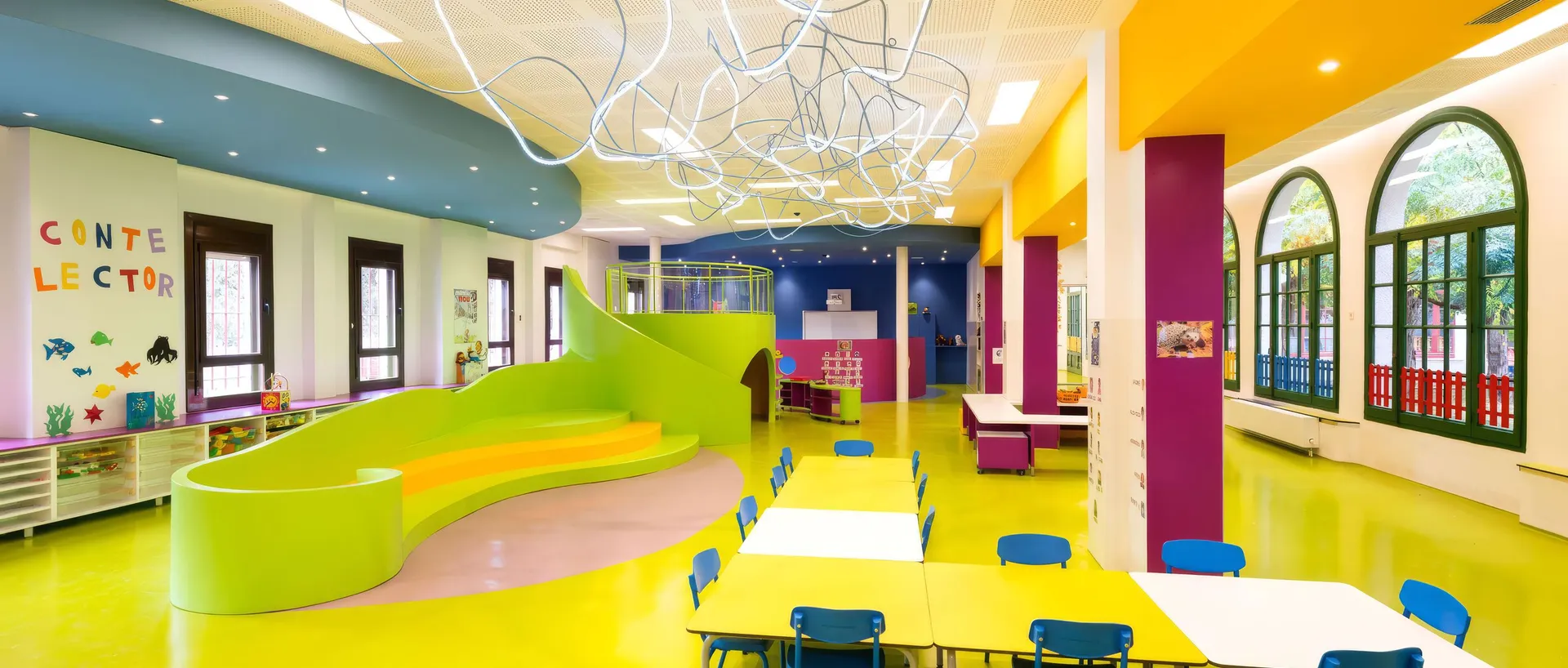 Espacio infantil de un colegio, sin personas: mesas amarillas con sillas azules, una grada verde ondulada con escalones, columnas de colores y un rótulo con el texto «Conte lector».