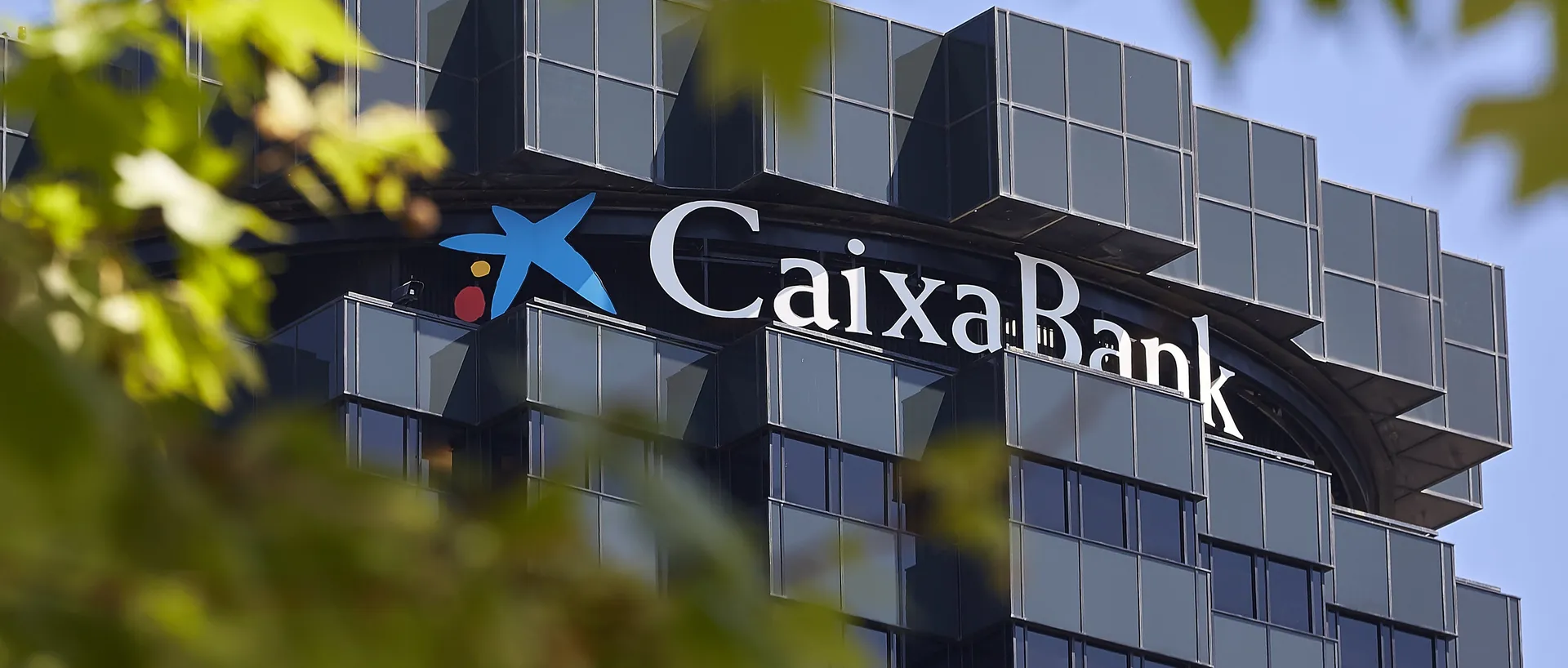 Lo alto de la torre de oficinas de CaixaBank, con el logotipo de la estrella azul y el nombre en letras blancas sobre la fachada de cristal oscuro, enmarcada por hojas de árbol desenfocadas en primer plano.