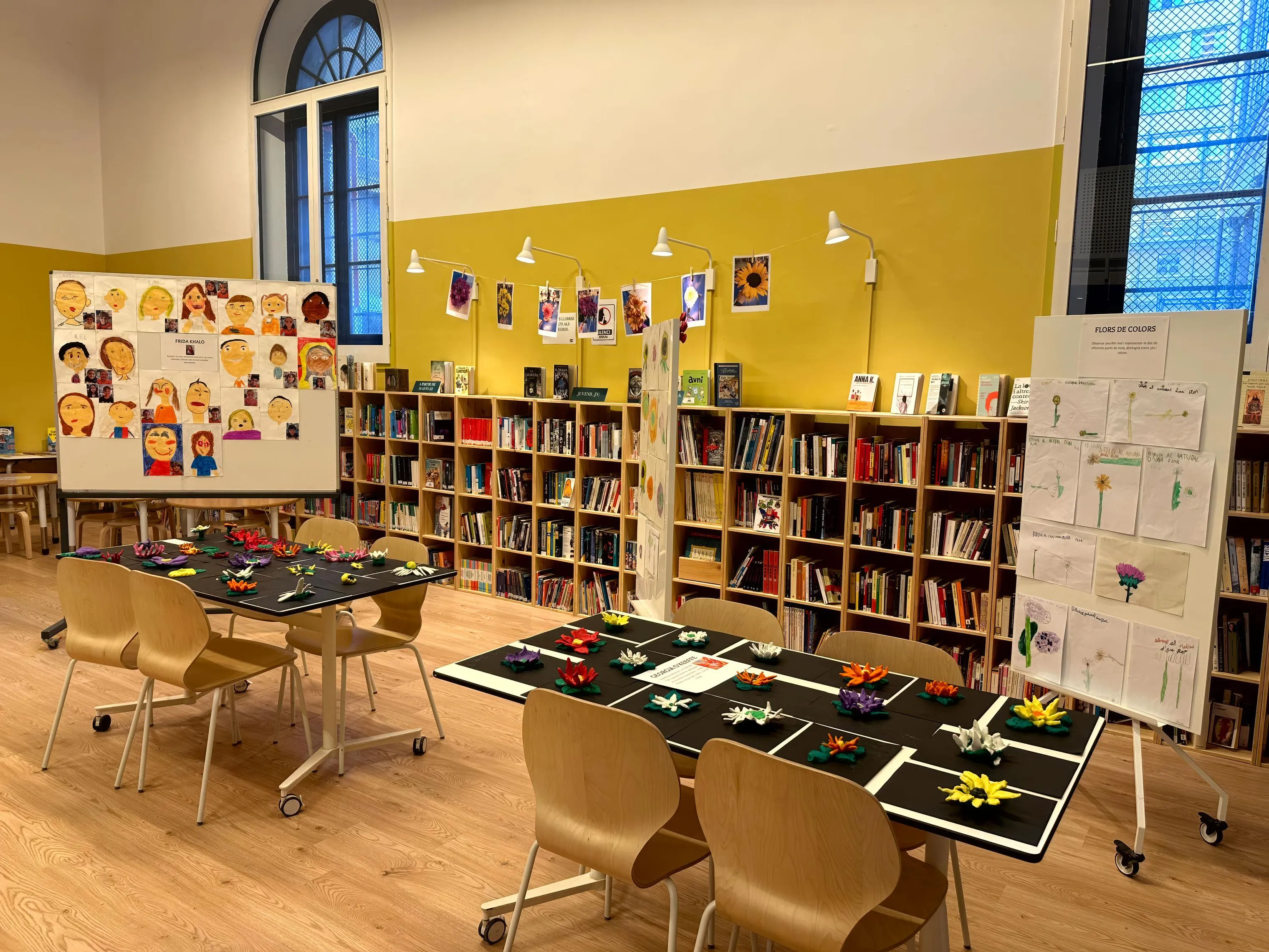 Biblioteca escolar sin personas, con estanterías llenas de libros, un panel de autorretratos infantiles junto a una ficha de Frida Kahlo y dos mesas con flores de plastilina de colores.